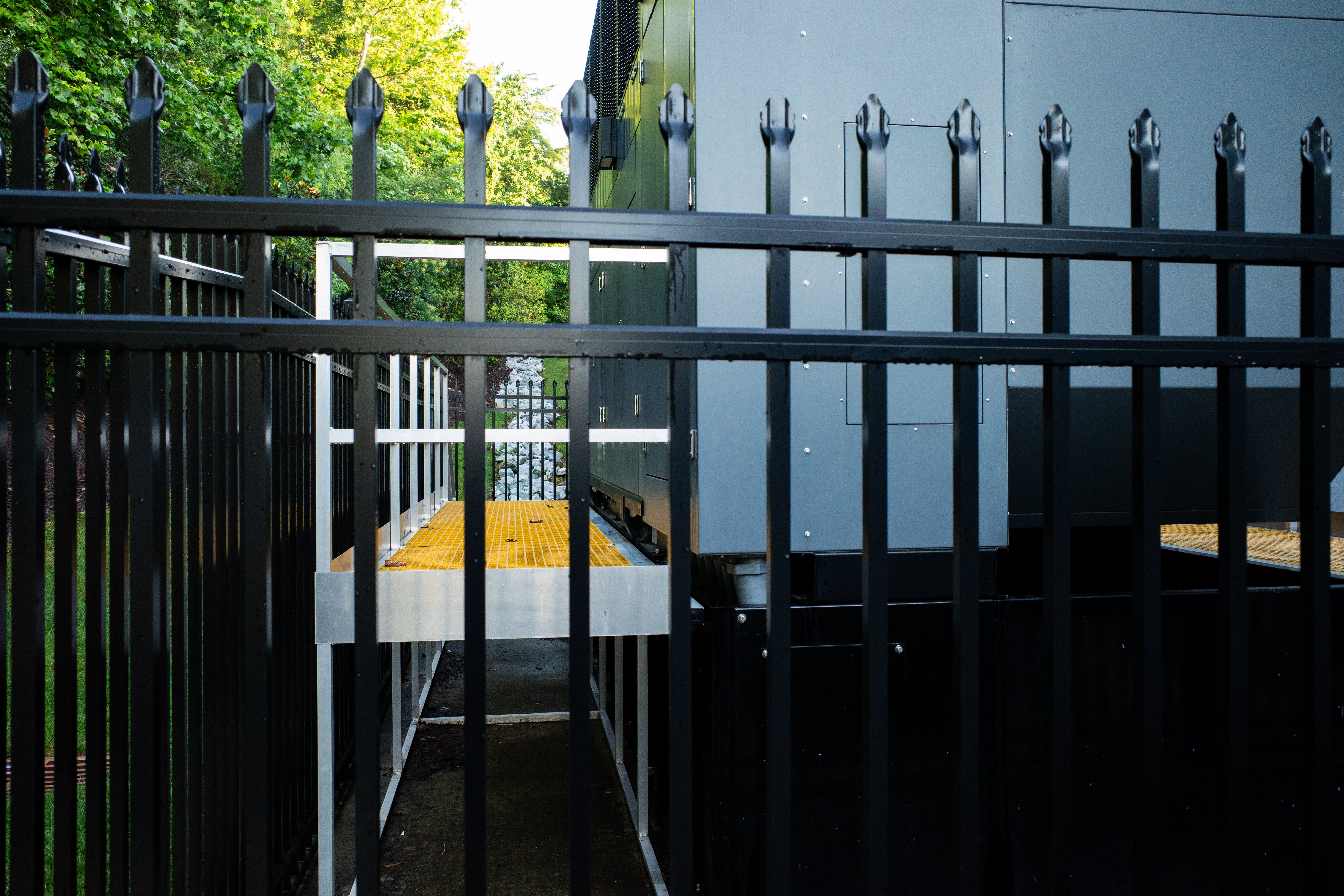 A color photo of a gate with many vertical bars and finials, a metal platform painted yellow and in the distance a stone watershed and foliage.