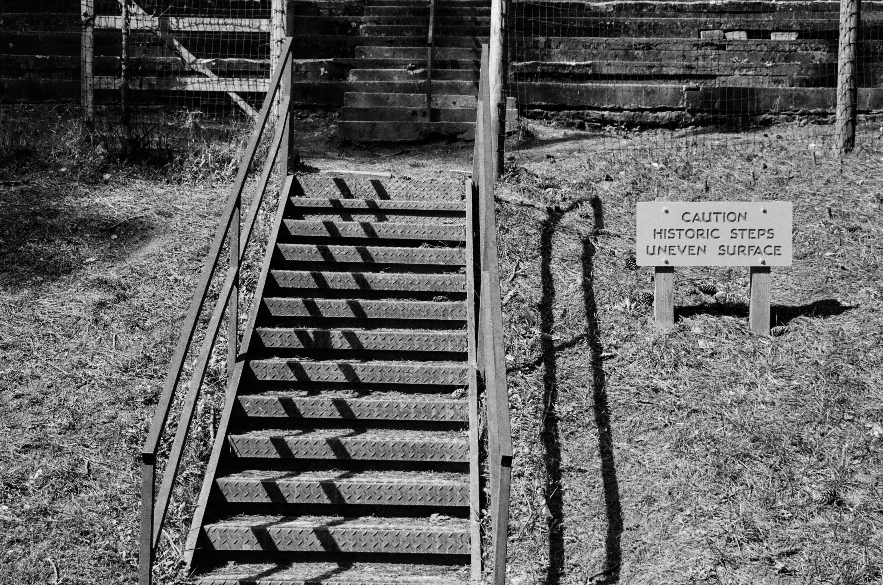 A black and white photo of a set of metal stairs followed by a set of concrete steps.  There is a sign to the right of the metal stairs saying, "Caution Historic Steps Uneven Surface".