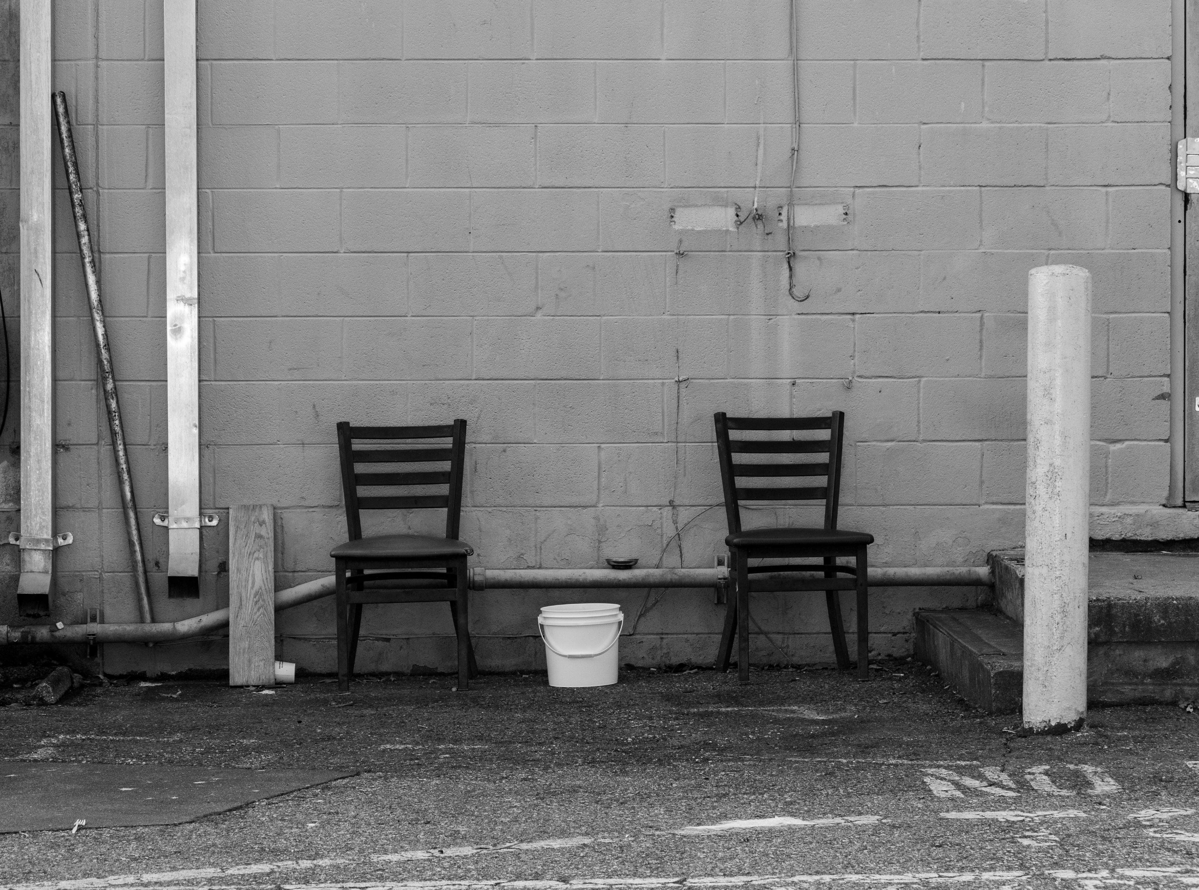 A black and white photo of the back of a strip mall building with two chairs with a bucket between them.