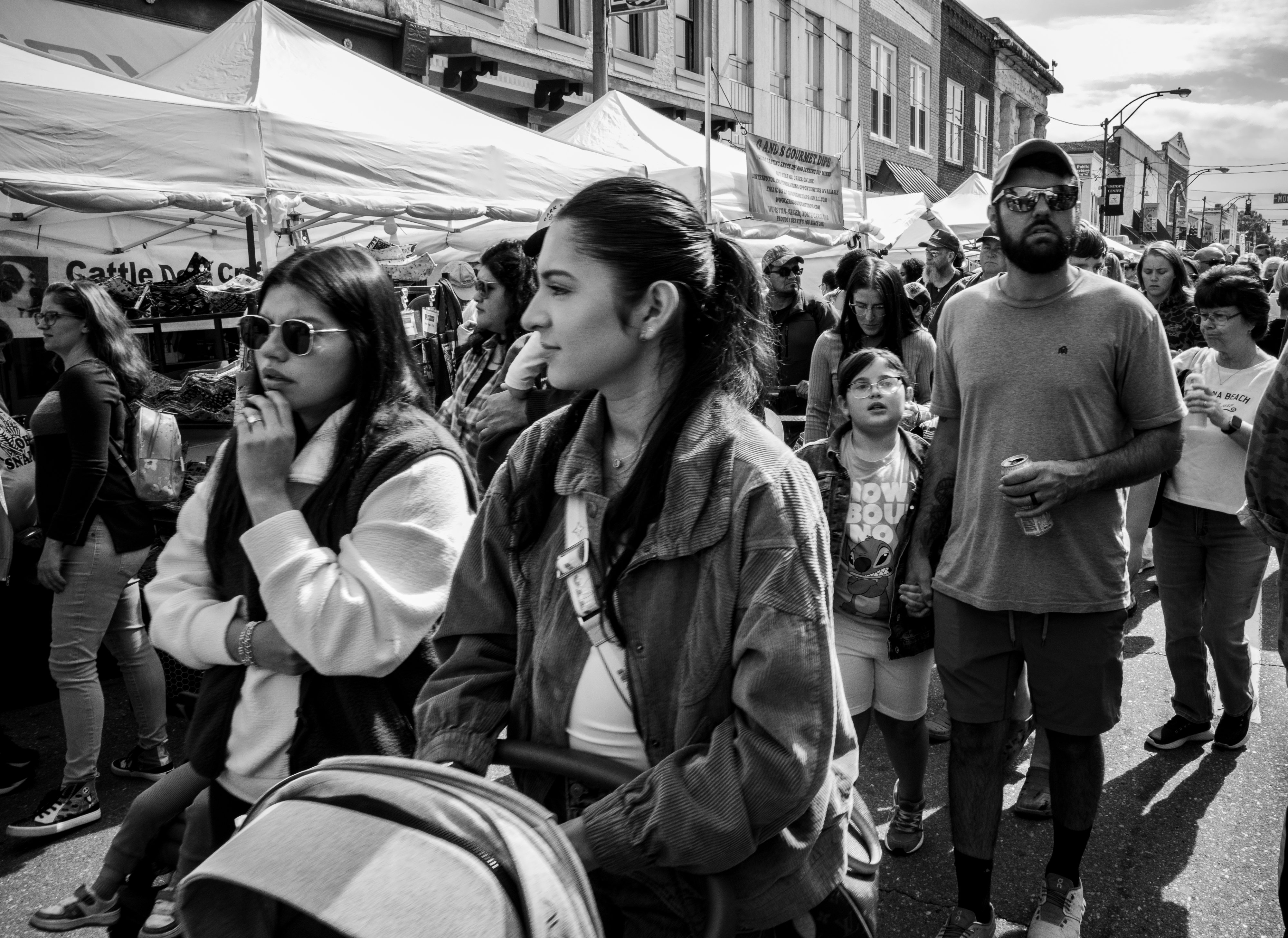 A black and white photo of a crowded street during a festival.  There are two women in the foreground, one pushing a barely seen buggy.  Just behind them is a man with a cap and sunglasses holding hands with his daughter.  All of this in the middle of a crowd of people on the street.