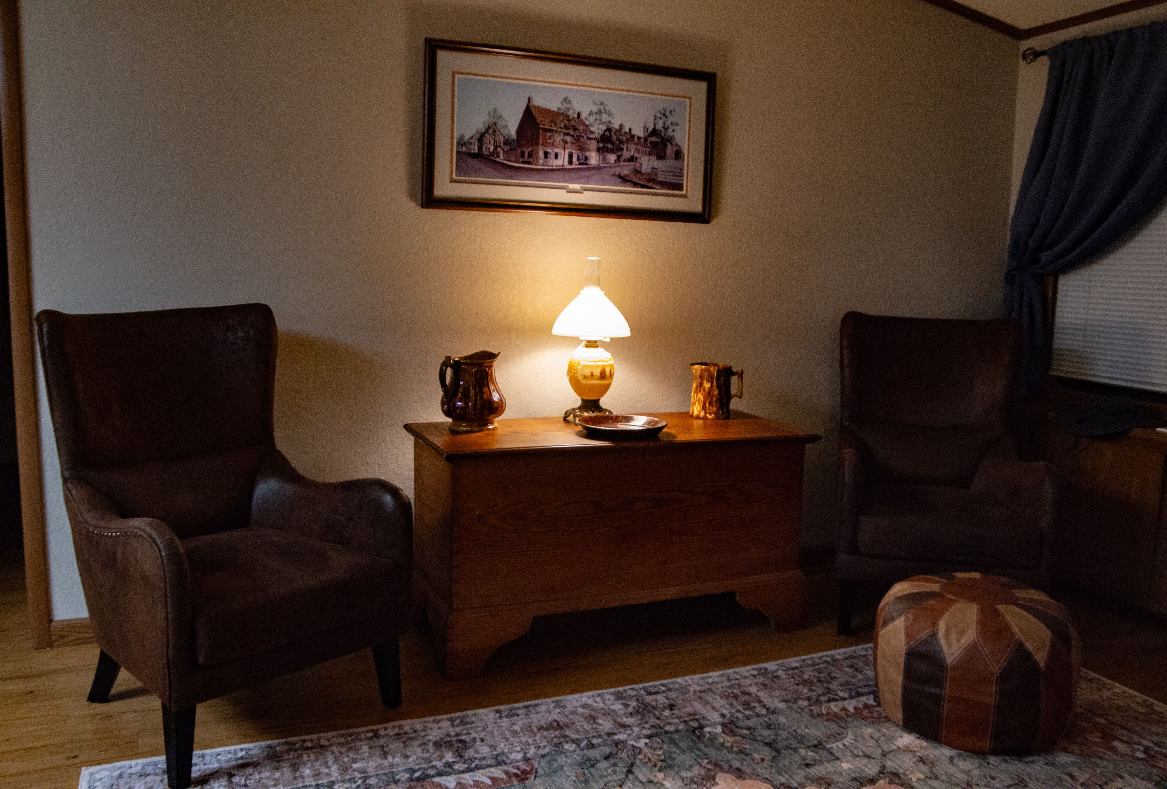 A color photo of the interior of house showing two matching chairs on either side of a large antique blanket chest.  There is a hassock in front of the right hand chair sitting on an oriental rug.  On the blanket chest there are two different mottled brown pitchers a deep plate and and oil lamp which has been converted to electric with a white glass shade.  There is a window to the far right with a closed shade and a curtain which has been pulled back.  On the wall above the blanket chest there is a rectangular painting of a 17th century house with streets, surrounding buildings and trees.