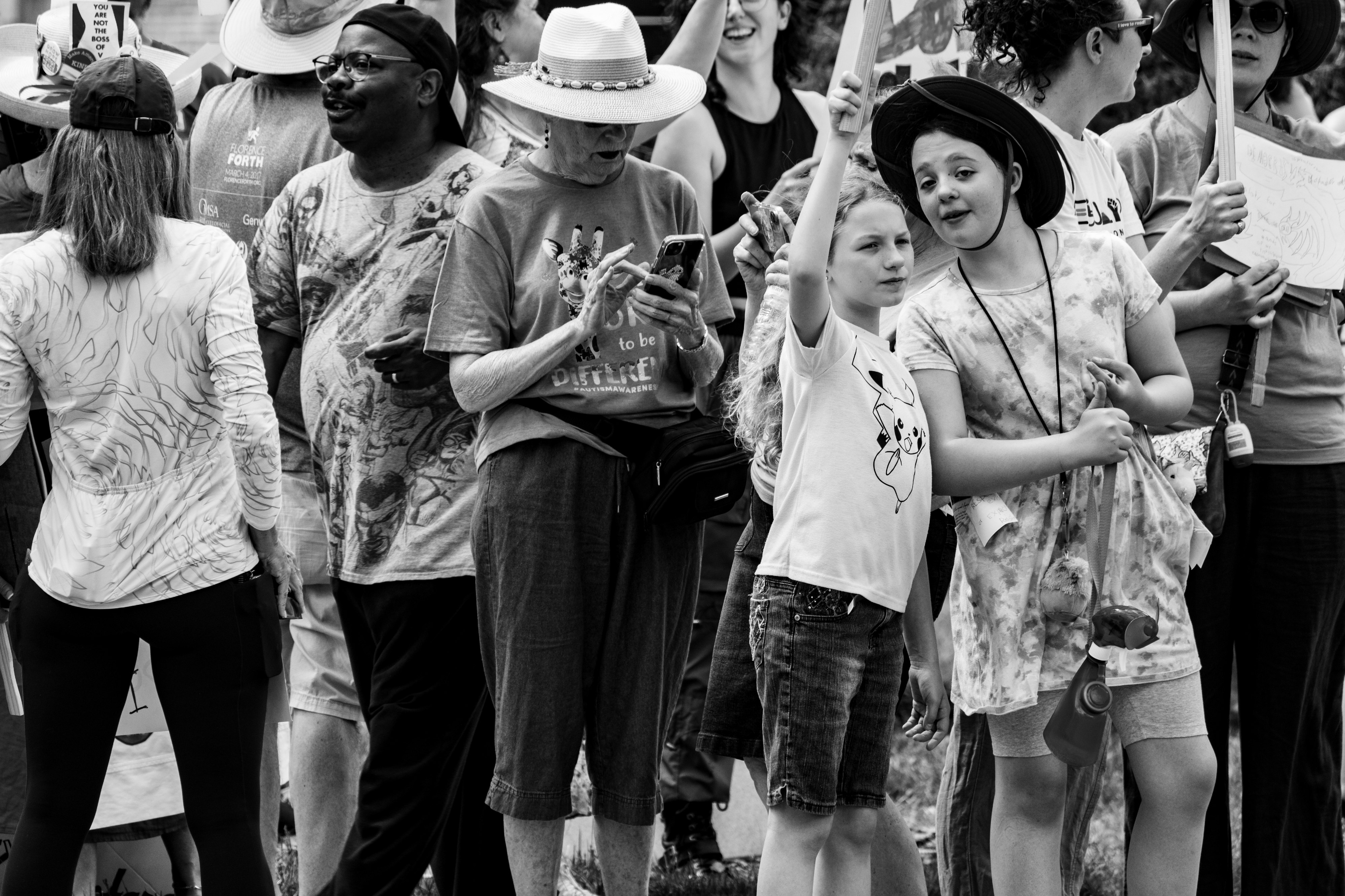 A black and white photo of a scene at a protest rally.  It is very crowded with people looking in every direction.  There is an elderly woman with a light colored wide brimmed hat on her phone in the center of the shot.