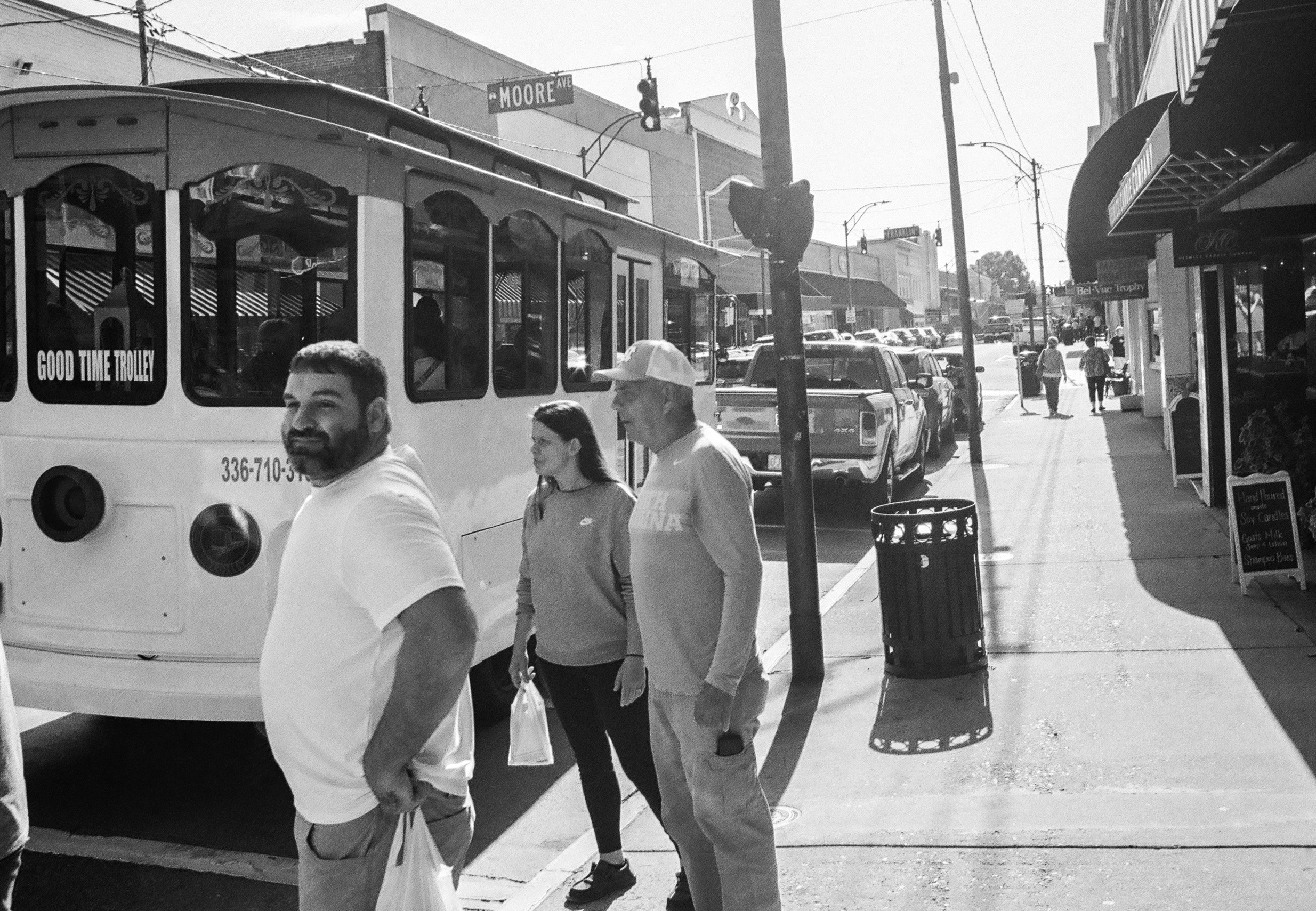 A black and white photo of a group of three people on a sidewalk facing the street where there is a trolley parked.