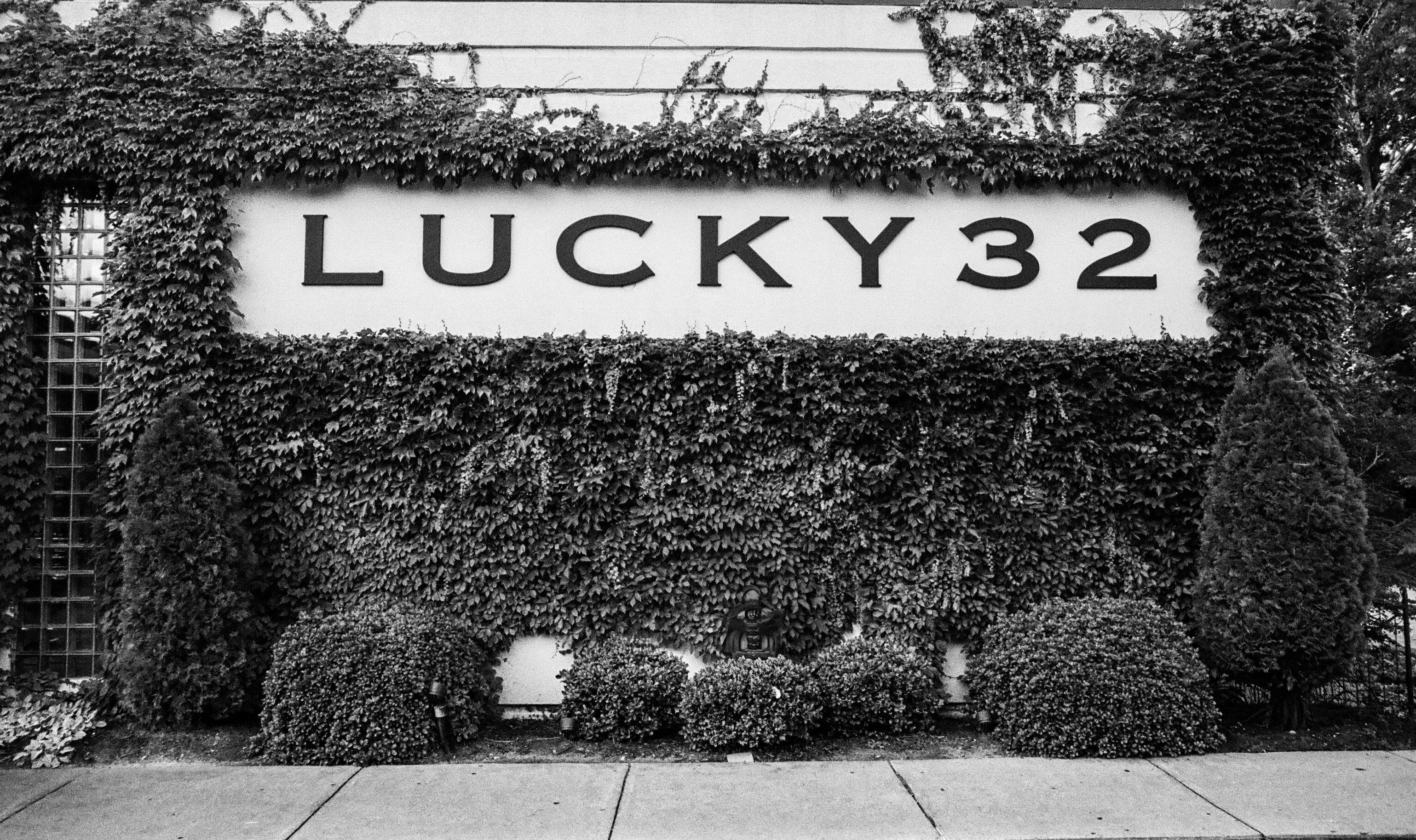 A black and white photo of a section of the outside wall of a restaurant called "Lucky 32".  The name is written in large font inside a rectangular cut out in the ivy growing up the side of the building.