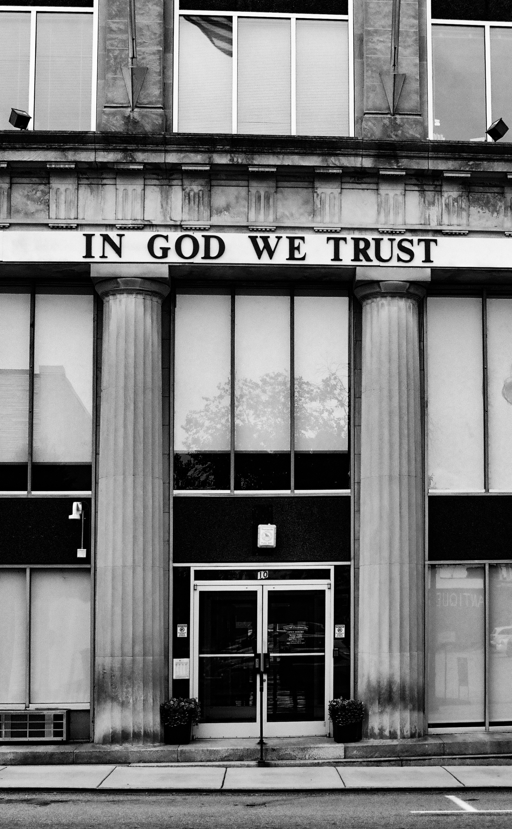 A black and white photo of the entrance to a government building in a small city.  There are columns on either side of the doors and the motto, "In God We Trust" over the entryway in large letters.
