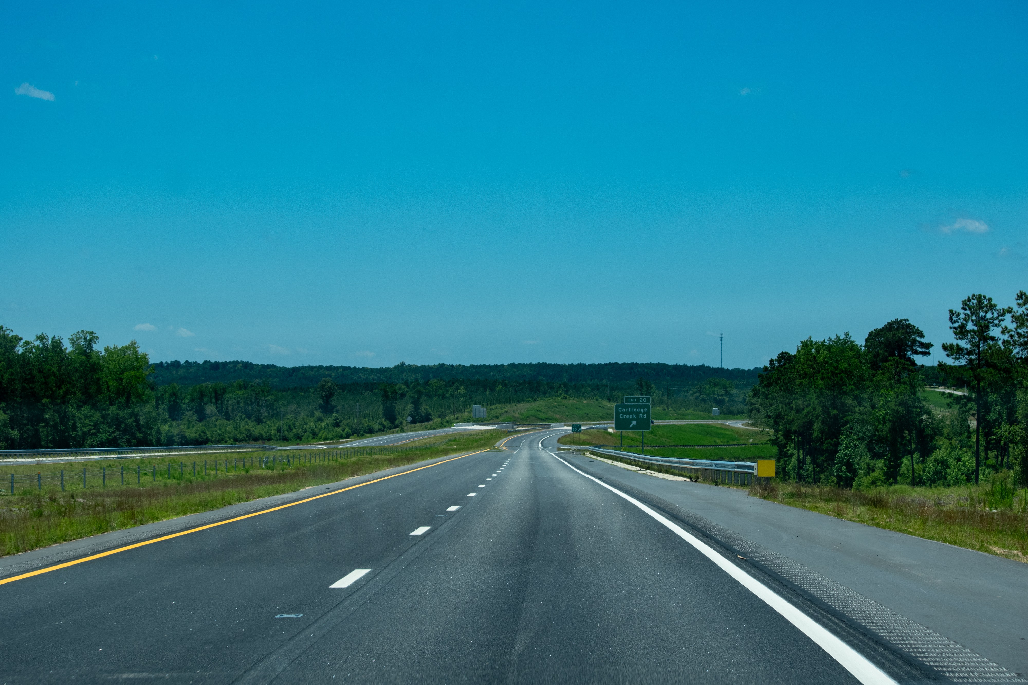 A color photo of a long stretch of highway blacktop heading off into a slow disappearing curve in the distance.  There is a wide grass median between the four lanes and forested hills in the background.