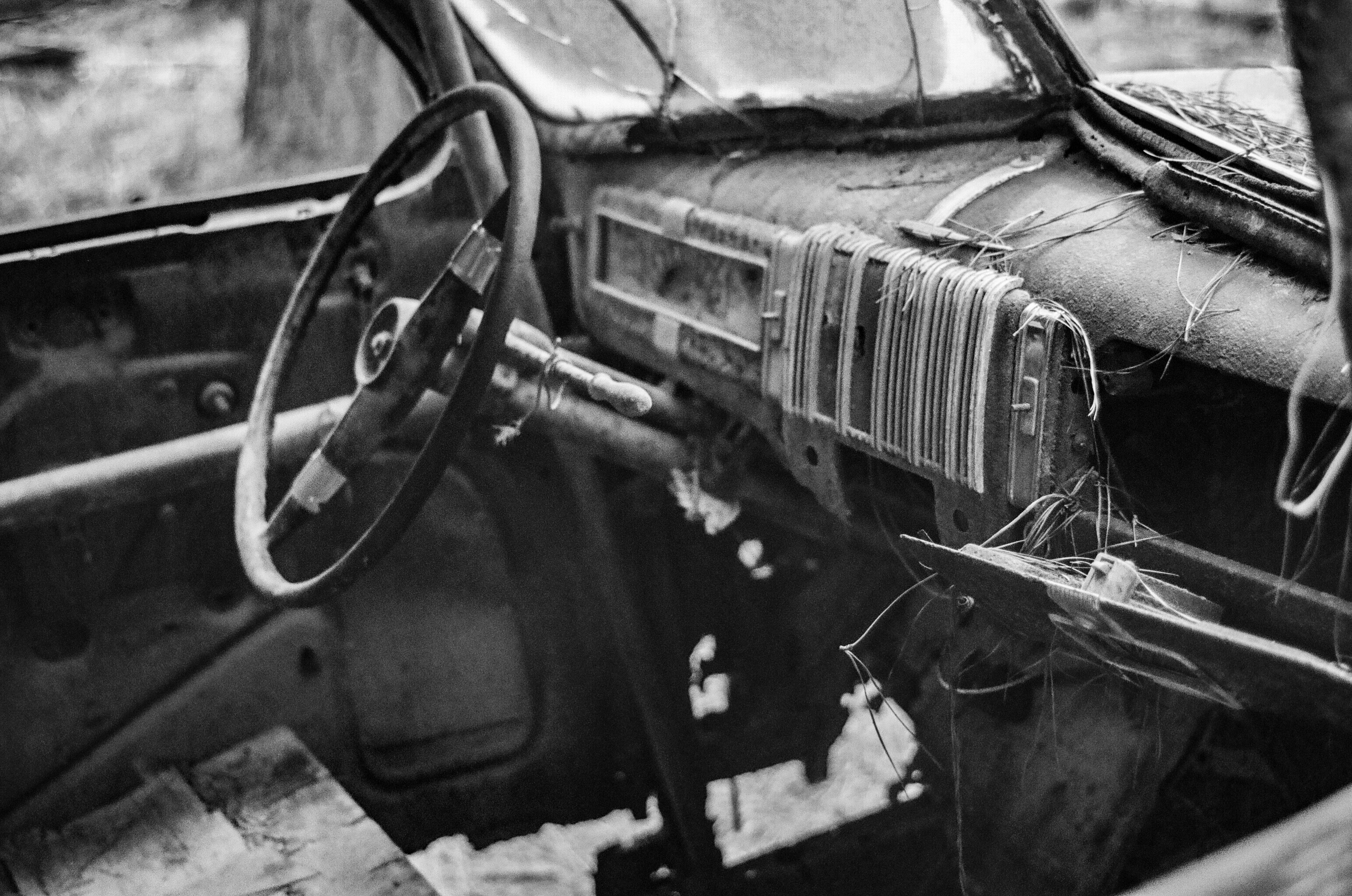 A black and white photo of the interior of a 1940s or '50s long abandoned race car showing the dashboard, steering wheel, driver's side door and floorboard.