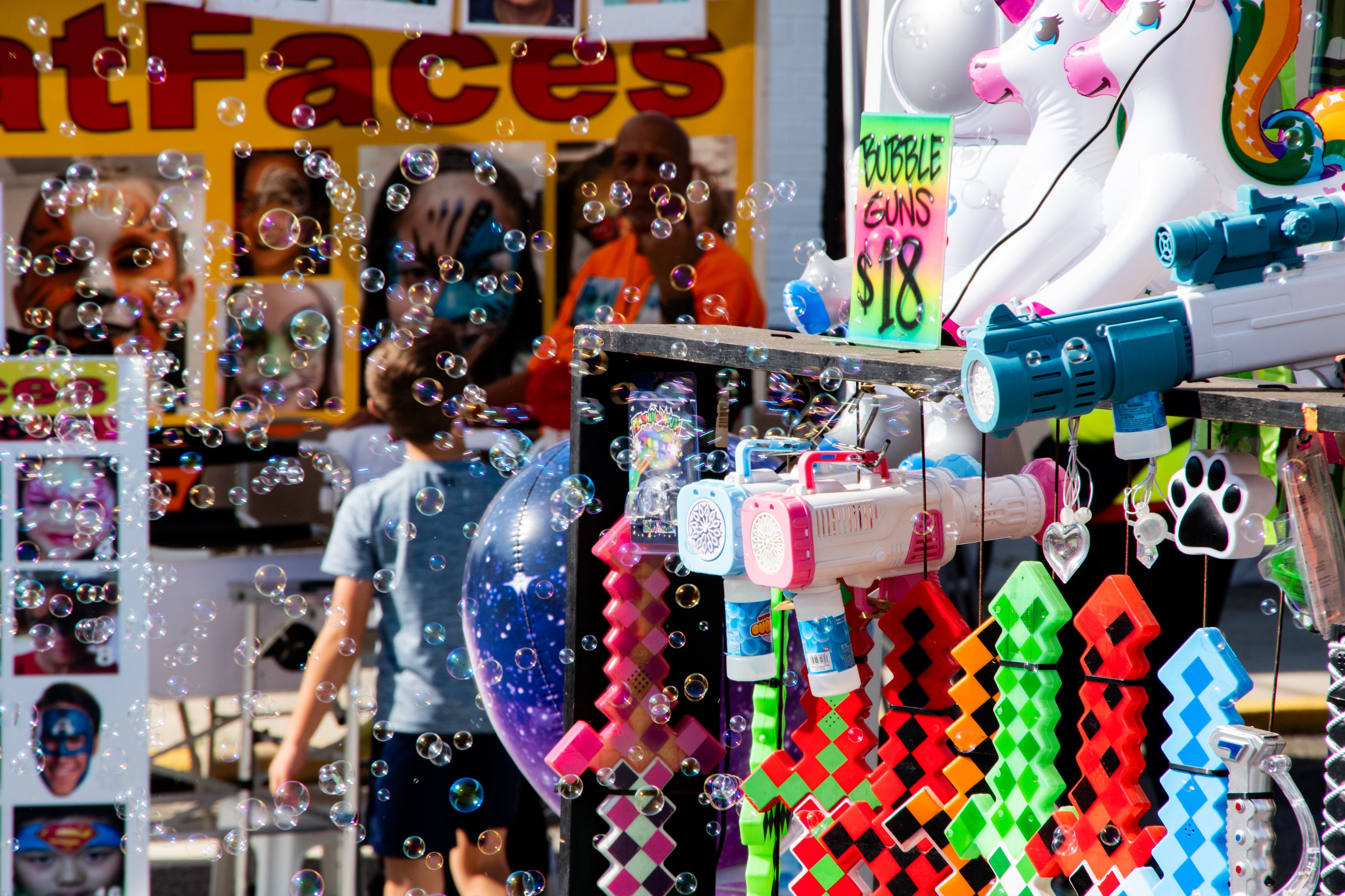 A color photo of the inside of a festival sales booth offering pixel swords, face painting, blow up unicorns and most prominently bubble guns which are active and filling the whole booth with bubbles.